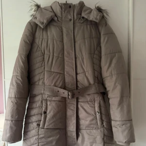 Beige lång dunjacka med bälte, strl 40 - Lång beige jacka med avtagbar huva och fuskpäls, bälte i midjan och snygga quiltade detaljer. Jackan har dragkedja och knappar framtill samt stora fickor. Perfekt för kalla dagar och ger en clean, stilren look. Använd sparsam. Bröst 108cm, längd 95cm.