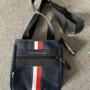 Blå axelväska från Tommy Hilfiger - Snygg blå axelväska från Tommy Hilfiger med justerbar svart rem. Endast anväd en gång, så i absolut nyskick. Nypris runt 800kr. Ett stort fack med dragkedja och två mindre i det stora. 