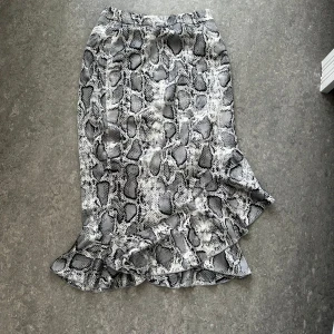 Grå långkjol med ormmönster från Missguided - Säljer en snygg långkjol från Missguided med grått och svart ormmönster. Kjolen har en volangkant nedtill och en diskret dragkedja i sidan. Perfekt för dig som vill sticka ut med ett coolt mönster och gillar trendiga detaljer.