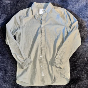 grön linneskjorta med knappar - Stilren grön skjorta från Arket i mjuk linne. Klassisk krage, långa ärmar och knäppning framtill. Perfekt att bära öppen över en t-shirt eller stängd för en clean look. Enkel och tidlös design som funkar till många outfits.