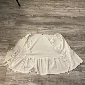 Vit omlottklänning från 157 - Supersöt vit omlottklänning från 157 med volangkant och knyt i midjan. Klänningen har en luftig känsla och är gjord i ett strukturerat bomullstyg. Perfekt för varma dagar och enkel att styla med sneakers eller sandaler.