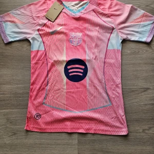 Barça Lamine Yamal rosa matchtröja Nike - Säljer en FC Barcelona fotbollströja från Nike med Lamine Yamal och nummer 10 på ryggen. Tröjan är rosa med ljusblå och vita detaljer, kortärmad och har tryck med Spotify-logga på bröstet. Materialet är lätt och andas, perfekt för fotbollsträning eller match.