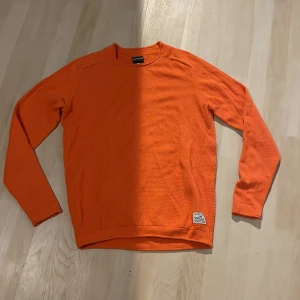 Orange långärmad tröja Jack & Jones - Snygg orange långärmad tröja från Jack & Jones. Tröjan har en diskret struktur på framsidan och en liten patch nertill. Perfekt för dig som gillar färg och vill sticka ut lite extra. Skön passform och rund halsringning.