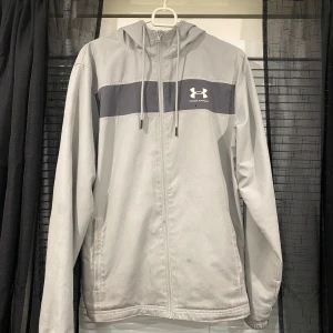 Ljusgrå vindjacka från Under Armour - Snygg ljusgrå vindjacka från Under Armour med mörkgrå panel över bröstet och vit logga. Jackan har huva med snörning, dragkedja framtill och två sidofickor. Perfekt för blåsiga dagar och sportiga outfits.