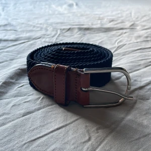 Svart flätat bälte från Saddler - Stilrent svart flätat bälte från Saddler med detaljer i brunt äkta läder och silvrigt spänne. Perfekt accessoar för att lyfta din outfit med en klassisk touch. Bältet har en snygg kombination av textil och läder.