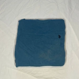 Blå stickad tröja från U.S. Polo Assn. - Snygg blå stickad tröja från U.S. Polo Assn. med klassisk rund halsringning och broderad logga på bröstet. Tröjan har långa ärmar och ribbade muddar vid ärmslut och nederkant. Perfekt för lager-på-lager och chill stil.