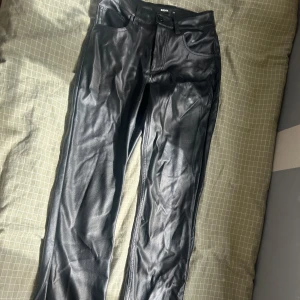 Svarta skinnbyxor från Bikbok - Snygga svarta byxor i skinnimitation från Bikbok. De har klassisk femficksdesign, raka ben och midjehällor för bälte. Materialet är glansigt och ger en cool look, perfekt för dig som vill sticka ut lite extra.