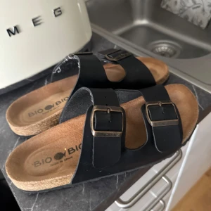 Svarta sandaler från Bio Bio - Svarta sandaler från Bio Bio med två breda remmar i skinn och justerbara metallspännen. Fotbädden är i naturfärgad mocka och sulan är i kork-look. Perfekta för en chill sommarstil.