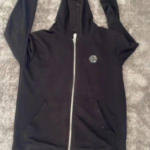 Svart hoodie från Philipp Plein - Svart hoodie från Philipp Plein med dragkedja framtill och klassisk huva. Tröjan har en snygg logga på bröstet och två stora fickor framtill. Tillverkad i mjuk bomullsmix som känns skön mot huden. Perfekt för en chill och stilren look.