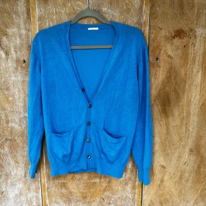 Blå kofta med knappar och fickor - Snygg blå kofta med v-ringning, knäppning framtill och två praktiska fickor. Långärmad och i mjukt stickat material, perfekt att slänga på sig över en t-shirt eller skjorta. Enkel och stilren design som funkar till många outfits.