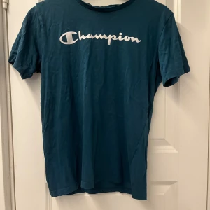 Blå Champion t-shirt med logga - Snygg blå t-shirt från Champion i storlek XL med klassisk vit logga tryckt över bröstet. T-shirten har rund halsringning och korta ärmar, perfekt för en avslappnad och sportig stil. Tillverkad i mjuk bomull som känns skön mot huden.