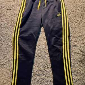 Adidas svarta träningsbyxor - Hej! Jag säljer ett par svarta Adidas träningsbyxor med tre gula ränder längs benen och Adidas-logga på låret. Byxorna har snörning i midjan och dragkedja vid höften. Tillverkade i Primegreen-material, vilket är återvunnen polyester. Perfekta för träning! Hör gärna av er vid funderingar!