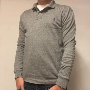 Grå långärmad pikétröja Polo Ralph Lauren - Säljer en klassisk grå långärmad pikétröja från Polo Ralph Lauren. Tröjan har krage, knappar vid halsen och den ikoniska lila broderade loggan på bröstet. Perfekt till jeans eller chinos för en clean och stilren look.  Nypris:1600
