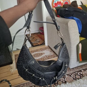 Svart DKNY axelväska med mönster - Snygg svart axelväska från DKNY med deras klassiska logomönster över hela väskan. Väskan har silverfärgade ringdetaljer, justerbar axelrem och dragkedja upptill. Materialet är en mix av skinn och textil, och framsidan har cool snörningsdetalj med metallringar.