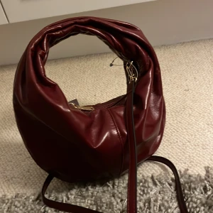 Vinröd axelväska i skinnimitation - Säljer en vinröd axelväska med rundad form och lång justerbar rem. Väskan är gjord i glansig skinnimitation och har guldfärgade metalldetaljer samt dragkedja upptill. Perfekt för dig som vill ha en snygg och trendig accessoar till din outfit. Helt ny, helt oanvänd! Alla etiketter sitter kvar.