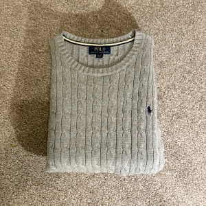 Grå kabelstickad tröja Polo Ralph Lauren - Säljer en grå kabelstickad tröja från Polo Ralph Lauren i storlek barn XL. I vuxen storlek är den S/M. Tröjan har rund halsringning, långa ärmar och den klassiska broderade loggan på bröstet.