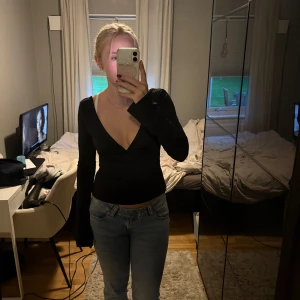 Svart långärmad omlottopp med v-ringning - Säljer en svart långärmad omlottopp med djup v-ringning framtill och öppen rygg med knyt i midjan. Toppens material känns mjukt och stretchigt, perfekt för att skapa en snygg siluett. Passar dig som gillar stilrena och trendiga plagg med lite edge.