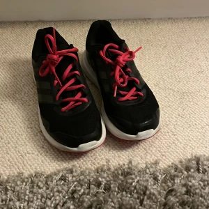 Svarta Adidas sneakers med röda snören - Säljer ett par svarta Adidas sneakers med klassiska tre ränder på sidan och röda snören. Skorna har vit sula och ovandel i mesh och syntet, vilket gör dem både lätta och bekväma. Perfekta för dig som gillar sportig stil och vill sticka ut med färgglada detaljer.