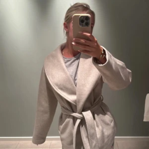 Beige omlottkappa med bälte - Snygg beige kappa med bred krage och omlottmodell. Jackan har ett matchande bälte i midjan som knyts och ger en schysst siluett. Långärmad och i ett mjukt, ulligt material som känns lyxigt och passar perfekt till lager-på-lager. 