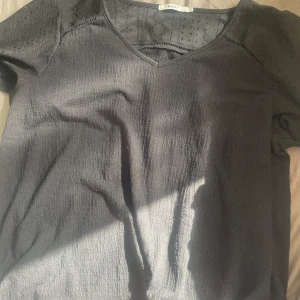 Svart blus med broderade detaljer &GOLF - Svart blus från &GOLF i storlek L med v-ringning och korta ärmar. Överdel och ärmar har broderade hålmönster och diskreta puffärmar. Tyget är lätt strukturerat och ger en cool look. Perfekt för dig som gillar stilrena men ändå unika plagg.