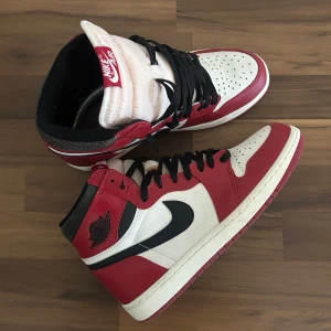 Nike Air Jordan 1 Lost and Found - Säljer ett par Nike Air Jordan 1 High i den klassiska Chicago-färgen. Skorna har röd och vit ovandel i skinn med svarta detaljer och svart swoosh. Svart snörning och ikonisk Air Jordan-logga på sidan. Perfekta för dig som vill sticka ut med en tidlös sneaker.