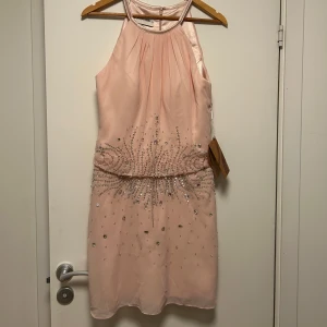 Ljusrosa kort klänning med paljetter - Säljer en ljusrosa kort klänning med halterneck och vackra paljettdetaljer 💗💗. Klänningen har ett tunt, skirt tyg och är ärmlös med en elegant, veckad överdel. Helt ny med prislappar kvar! Priset går att diskutera 