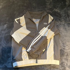 Ascend vindjacka i grått och beige - Snygg vindjacka från Ascend i grått och beige med coola blockmönster och vita ränder längs ärmarna. Jackan har dragkedja framtill, hög krage och elastiska muddar. Perfekt för dig som gillar sportig och retro stil.