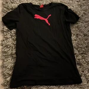 Svart Puma t-shirt med röd logga - Säljer en svart t-shirt från Puma i storlek 34/36. T-shirten har en klassisk passform med korta ärmar och en stor röd Puma-logga på bröstet. Perfekt för dig som gillar sportiga och stilrena plagg. Materialet känns mjukt och bekvämt.