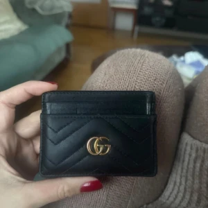 Svart kortfodral från Gucci i skinn - Supersnyggt svart kortfodral från Gucci i äkta skinn med klassiskt GG-logo i guld framtill. Fodralet har flera kortfack och ett stilrent, quiltat mönster som ger en lyxig känsla. Perfekt för dig som vill ha koll på korten med stil.