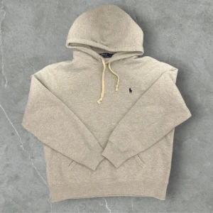Grå hoodie från Polo Ralph Lauren - Snygg grå hoodie från Polo Ralph Lauren med klassisk broderad logga på bröstet. Tröjan har huva med snörning, känguruficka och ribbade muddar.  Inga defekter eller dåliga saker med tröjan. Sitter som M/L