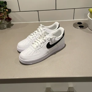 Nike Air Force 1 vita med svart swoosh - Klassiska Nike Air Force 1 sneakers i vitt skinn med svart swoosh. Lågt skaft, perforerad tå och vit platt sula. Perfekta för dig som gillar en clean och tidlös look. Skorna har snörning och är riktigt bekväma för vardagsbruk. Skorna är även helt oanvända och i storlek 44.