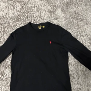 Svart långärmad tröja Polo Ralph Lauren - Svart långärmad tröja från Polo Ralph Lauren i klassisk fit. Tröjan har rund hals och är gjord i mjuk bomull. På bröstet finns den ikoniska röda Polo-loggan broderad. Perfekt för dig som gillar stilrena och tidlösa plagg.