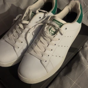 Adidas Stan Smith vita sneakers strl 44 - Klassiska Adidas Stan Smith sneakers i vitt skinn med gröna detaljer på hälen och tungan. Perfekt för dig som gillar en clean och tidlös look. Ikoniskt Stan Smith-ansikte på plösen. Skicket är 9/10 då sulan är smutsig men inte mer än det!