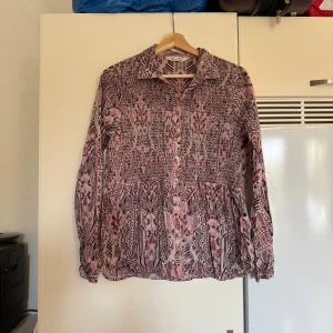 Mönstrad skjorta från Odd Molly - Supersnygg långärmad skjorta från Odd Molly med unikt mönster i rosa och vinröda toner. Skjortan har knappar framtill, krage och lätt rynkad nederdel. Perfekt för dig som gillar färg och detaljer. Materialet känns mjukt och luftigt. Storlek 2 som enligt deras storleksguide motsvarar en storlek M.  Mycket fint skick, jag hittar inga synliga anmärkningar eller defekter.