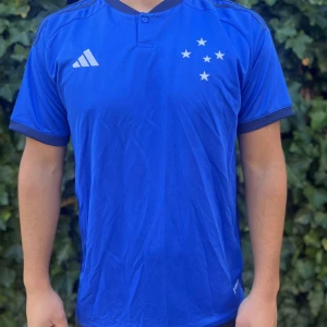 Blå Cruzeiro fotbollströja Adidas - Snygg blå fotbollströja från Adidas med Cruzeiro-stjärnor på bröstet och vit Adidas-logga. Kortärmad modell med mörkblå detaljer vid krage och ärmslut. Tillverkad i lätt och ventilerande material, perfekt för träning eller match. Nypris:599kr GORILLAPRICE:279kr																																											SKICK:10/10 med tagg kvar                    KVITTO MEDKOMMER