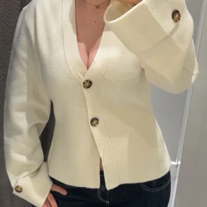 Vit cardigan jacka med knappar - Snygg vit cardigan jacka med stora bruna knappar och v-ringad hals. Jackan har lång ärm med knappdetalj vid ärmslut och en figurnära passform. Perfekt för dig som gillar stilrena och trendiga plagg.