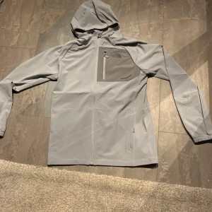 The North face windbreaker - Snygg ljusgrå vindjacka från The North Face med huva och dragkedja framtill. Jackan har en praktisk bröstficka med dragkedja och elastiska muddar vid ärmsluten. Perfekt för blåsiga dagar och enkel att matcha med det mesta.