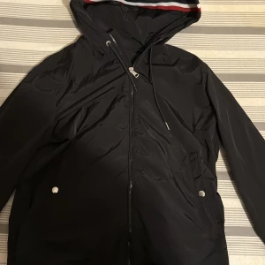 Svart vindjacka från Moncler - Snygg svart vindjacka från Moncler med huva och dragkedja framtill. Jackan har två fickor med knappar och en huva med röd, vit och blå rand. Perfekt för blåsiga dagar och enkel att matcha med det mesta.