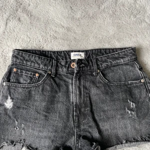 Svarta jeansshorts från ONLY - Säljer ett par svarta jeansshorts från ONLY med slitna detaljer och råa kanter. Klassisk femficksmodell med knapp och dragkedja framtill. Perfekta för en avslappnad stil.