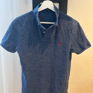 Blå piké från Polo Ralph Lauren - Säljer en blå pikétröja från Polo Ralph Lauren i slim fit-modell. Klassisk design med krage, knappar och den ikoniska röda loggan broderad på bröstet. Perfekt för en stilren och avslappnad look.