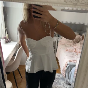 Vit korsett-topp med tunna axelband - Säljer en vit korsett-topp med tunna axelband och markerade kupor. Toppen har en figurnära passform upptill och faller ut lätt nedtill. Perfekt för att styla med jeans eller kjol.