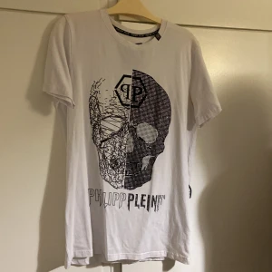 Vit t-shirt med döskalletryck från Philipp Plein - Säljer en vit t-shirt från Philipp Plein med ett stort svart döskalletryck framtill och logga på kragen. T-shirten har normal passform och är kortärmad. Perfekt för dig som gillar streetwear och unika prints.