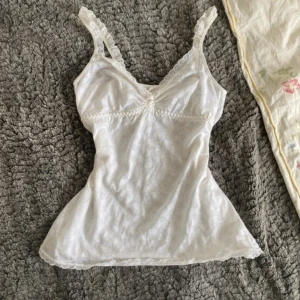 Vit spetslinnetopp - Säljer en söt vit linnetopp med spetsdetaljer längs axelbanden och nederkanten. Toppen har en feminin och romantisk känsla, perfekt att styla till olika outfits. Mjukt och lätt material.