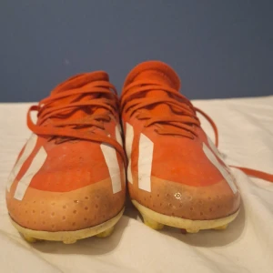 Röda fotbollsskor från Adidas - Säljer ett par röda fotbollsskor från Adidas med vita ränder och snörning. Skorna har en högre strumpliknande krage och platt sula med dobbar för bra grepp på planen. Perfekta för dig som vill sticka ut på fotbollsplanen!