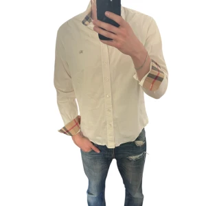 Vit skjorta med rutiga detaljer från Burberry - Säljer en klassisk vit skjorta från Burberry med ikoniska rutiga detaljer på krage och ärmslut. Skjortan har knappar framtill och lång ärm, perfekt för en stilren look. Storlek L. Är 185cm lång.