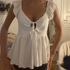 Vit blus med volangärmar och knytning - Söt vit blus med volangärmar och knytning framtill. Blusen har en lätt v-ringning och är tillverkad i ett luftigt material, perfekt för varmare dagar. Den har en lös passform nedtill och fina detaljer vid axlarna.