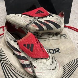 Adidas Predator fotbollsskor med röda detaljer - Säljer ett par Adidas Predator fotbollsskor i vitt med svarta och röda detaljer. Skorna har klassiska tre ränder på sidan, röda skyddslappar över snörningen och mönstrad ovandel. Perfekta för dig som vill sticka ut på planen.