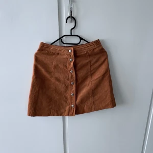 Brun kjol med knappar från H&M Divided - Kort brun kjol från H&M Divided med knappar framtill. Kjolen har en rak passform och är tillverkad i ett mjukt material som känns bekvämt. Perfekt att matcha med en enkel topp eller stickad tröja.