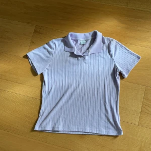 Ljuslila ribbad pikétröja från Lindex - Säljer en ljuslila ribbad pikétröja från Lindex med klassisk krage och korta ärmar. Tröjan har diskreta vertikala ränder i materialet och är perfekt för en avslappnad stil.