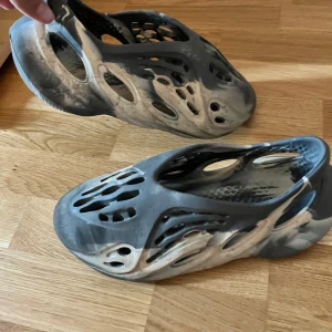 Grå och vita foam sandaler - Säljer ett par grå och vita sandaler i foam-stil med futuristisk design och många hål för ventilation. Skorna är tillverkade i syntetmaterial och har en rund tå samt platt sula. Perfekta för sommaren och riktigt lätta på foten.